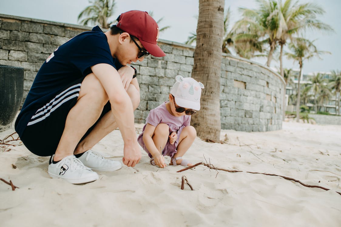 Free Full body of positive young Asian man wearing red cap playing with child on sandy shore with palms on background on clear day Stock Photo